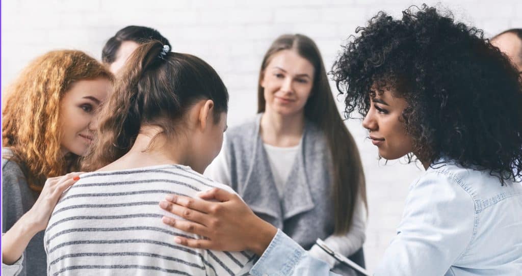 In an atmosphere reminiscent of a supportive communal environment, a group of women stands together. One woman gently comforts another by placing a hand on her shoulder, echoing the compassion needed for survivors of issues like those at the Arizona Juvenile Residential Treatment Center.